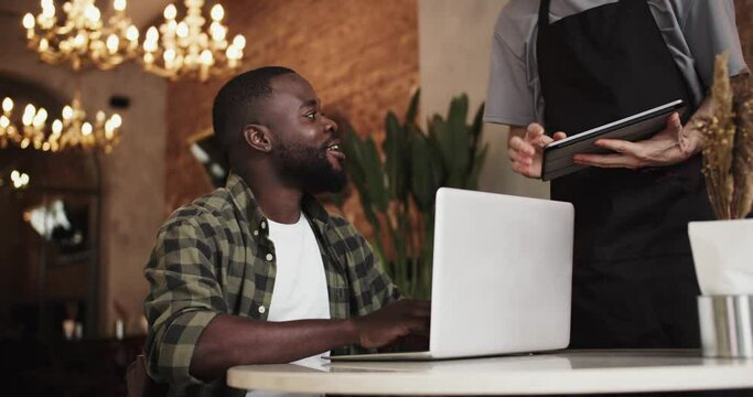 A black person with a beard works on a laptop in a cafe, a waiter approaches them and takes an order