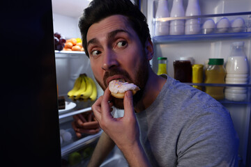 Man eating donut near refrigerator in kitchen at night. Bad habit