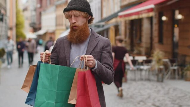 Happy smiling young adult man shopaholic consumer after shopping sale with full bags with gifts outdoors. Redhead bearded guy tourist traveler walking along the urban city street road. Town lifestyles