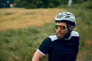Close-up portrait of young, serious,bearded man, cyclist in helmet and special glasses standing over filed, grass background. Concept of sport, hobby, leisure activity, training, health, endurance, ad