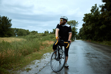 Fototapeta premium Sportive, young, bearded man, cyclist in uniform, helmet and glasses posing on bike, riding on road in chill evening. Concept of sport, hobby, leisure activity, training, health, speed, endurance, ad