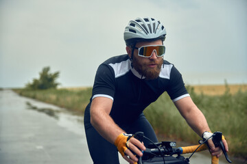 Young bearded man, cyclist in helmet, glasses and uniform riding bike on wet road in cloudy chill...