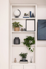 Fototapeta na wymiar Interior design. Shelves with stylish accessories, potted plants and frame near white wall