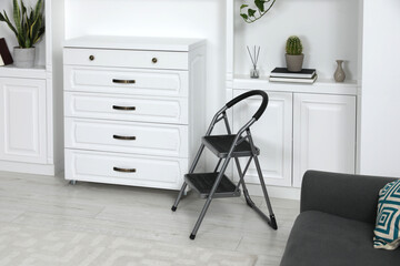 Metal folding ladder near chest of drawers and shelves with accessories in room