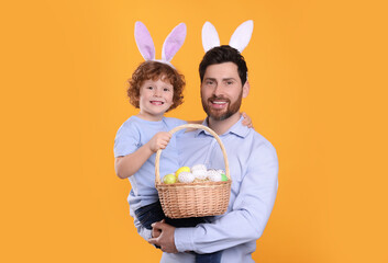 Happy father and son wearing cute bunny ears headbands on orange background. Boy holding Easter basket with painted eggs
