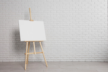 Wooden easel with blank canvas near white brick wall indoors. Space for text