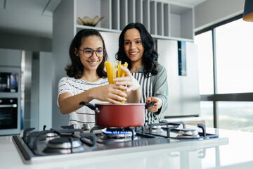 A mother and her teenage daughter joyfully cooking together in the kitchen, sharing laughter and...