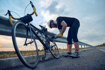 Fototapeta na wymiar Young bearded man, cyclist in sportswear and helmet, pumping up bicycle wheel on road. Riding in chill evening. Concept of sport, hobby, leisure activity, training, health, speed, endurance, ad