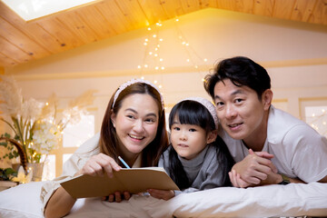 Happy family with mother and father lying teaching homework with daughter on bed in bedroom at...