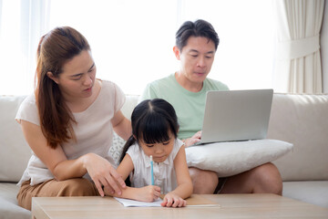 Happy family with mother teaching homework with daughter and father working with laptop on sofa in living room at home, mom explaining schoolwork with kid together, lifestyles and education.