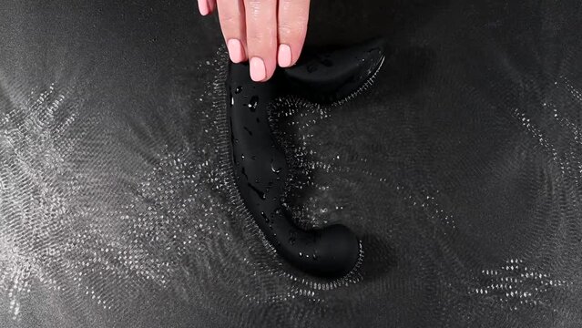 A woman holds a black prostate stimulator that vibrates and creates ripples on the surface of the water against a black background.