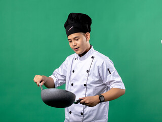 A portrait of an Indonesian Asian man wearing a chef's uniform and a chef's hat, posing with great joy, hands raised up holding a spatula and a frying pan, isolated with a green background.