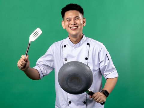 A portrait of an Indonesian Asian man wearing a chef's uniform, posing with great joy, hands raised up holding a spatula and a frying pan, isolated with a green background.