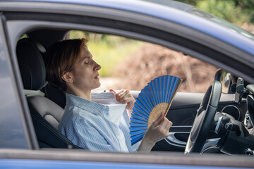 Tired exhausted middle aged woman waving blue fan suffers from stuffiness driving car on summer hot weather. Overheating, sultriness, high temperature, swelter in car with broken air conditioner. - obrazy, fototapety, plakaty