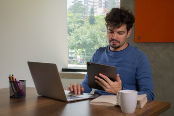 A focused young Latin man is working using a digital tablet at a casual workspace office.