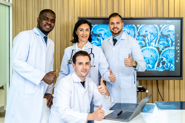 Group of medical practitioners in lab coats gesturing thumbs up while sitting and standing with smiling on camera. Four colleagues discusses the results of an MRI scan of the patient's head