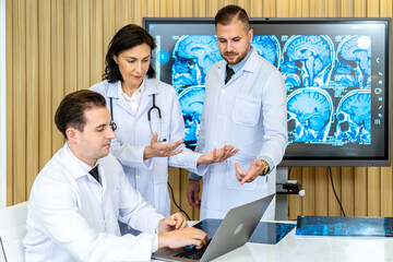 Fototapeta premium Team of three doctors discusses the results of an MRI scan of the patient's head in the consulting room of modern hospital, physician discussing about treatment for patient looking in laptop