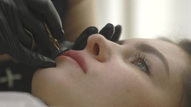 Young pretty woman patiently lying on the procedure of permanent lip makeup. A tattoo artist in black gloves gently applies strokes to the client's delicate lips