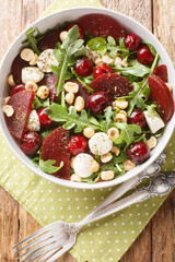 Fresh summer salad of arugula, baked beets, cherries, mozzarella, hazelnuts close-up in a bowl on the table. Vertical top view from above