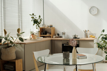 Part of spacious kitchen with round glass table with vases and notepads standing in the center against green domestic plants and counter