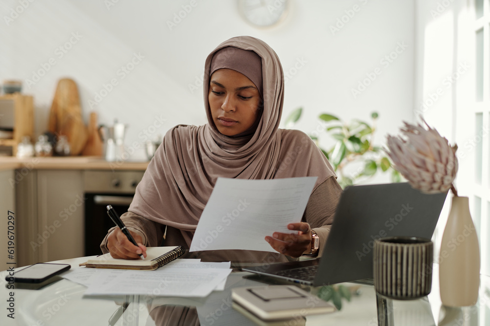 Poster young serious female auditor in hijab making notes in copybook while sitting by workplace and analyz