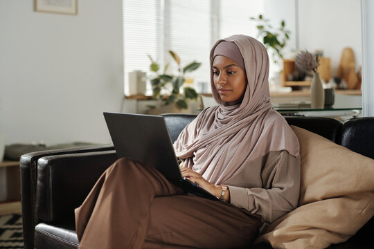 Young pretty African American woman in hijab typing on laptop keyboard while sitting on couch in living room and working over new project