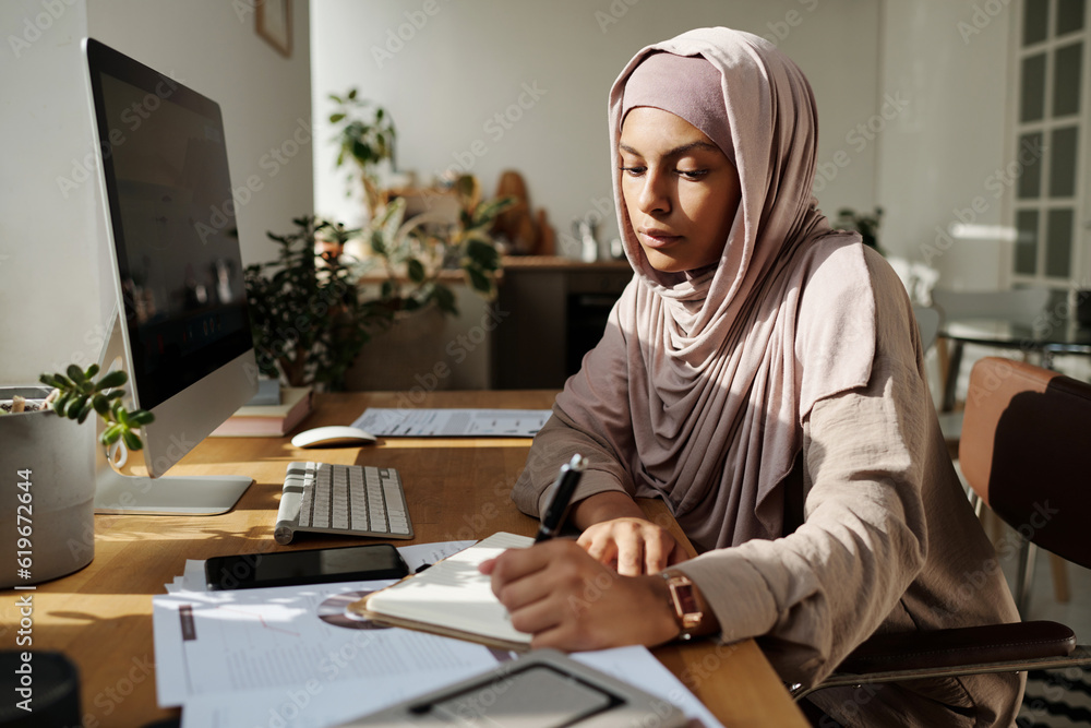 Poster Young serious Muslim businesswoman making notes in notepad ordocument while sitting by workplace in front of computer screen