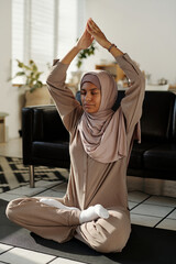Fototapeta premium Young serene Muslim woman in hijab and activewear sitting in pose of lotus with her hands put together over head and keeping eyes closed