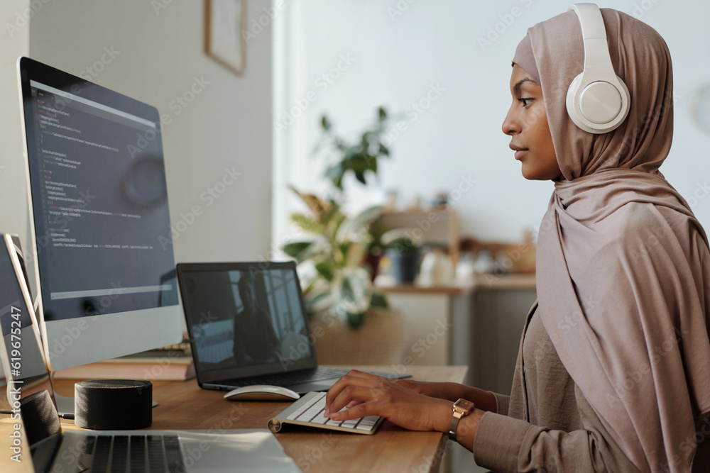 Aufkleber side view of young female programmer in headphones and hijab decoding online data while communicatin