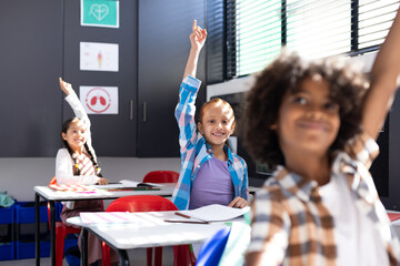 Diverse, happy children sitting at desks raising hands in elementary school classroom, copy space