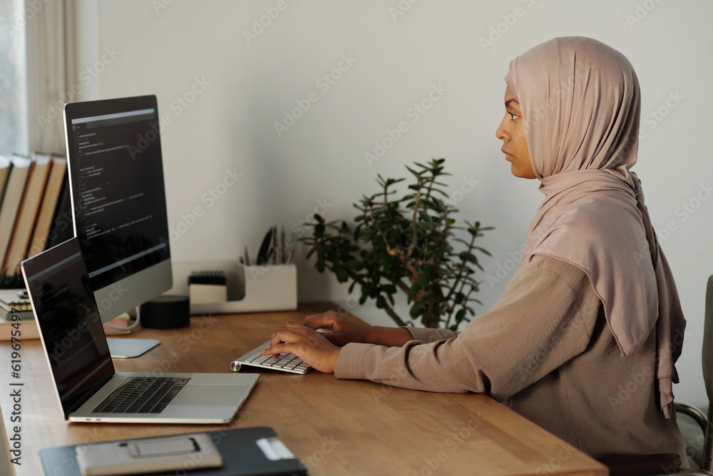 Poster young muslim businesswoman in hijab decoding data on computer screen and typing while sitting by wor