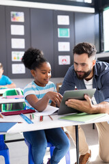 Vertical of happy male teacher and schoolgirl using tablet at her desk in classroom, copy space