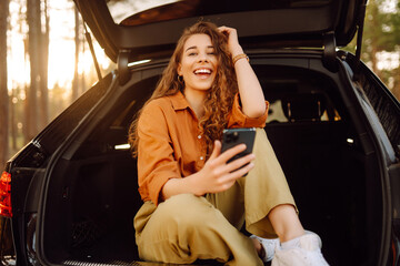 Naklejka premium Beautiful woman sits in the trunk of a car in the forest and uses a smartphone. The concept of traveling by car, active lifestyle.