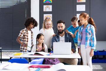 Diverse elementary schoolchildren around male teacher using laptop at desk in classroom, copy space