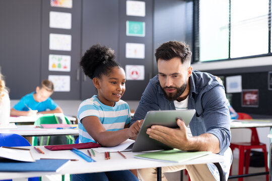 Diverse male teacher and happy girl using tablet at her desk in elementary classroom