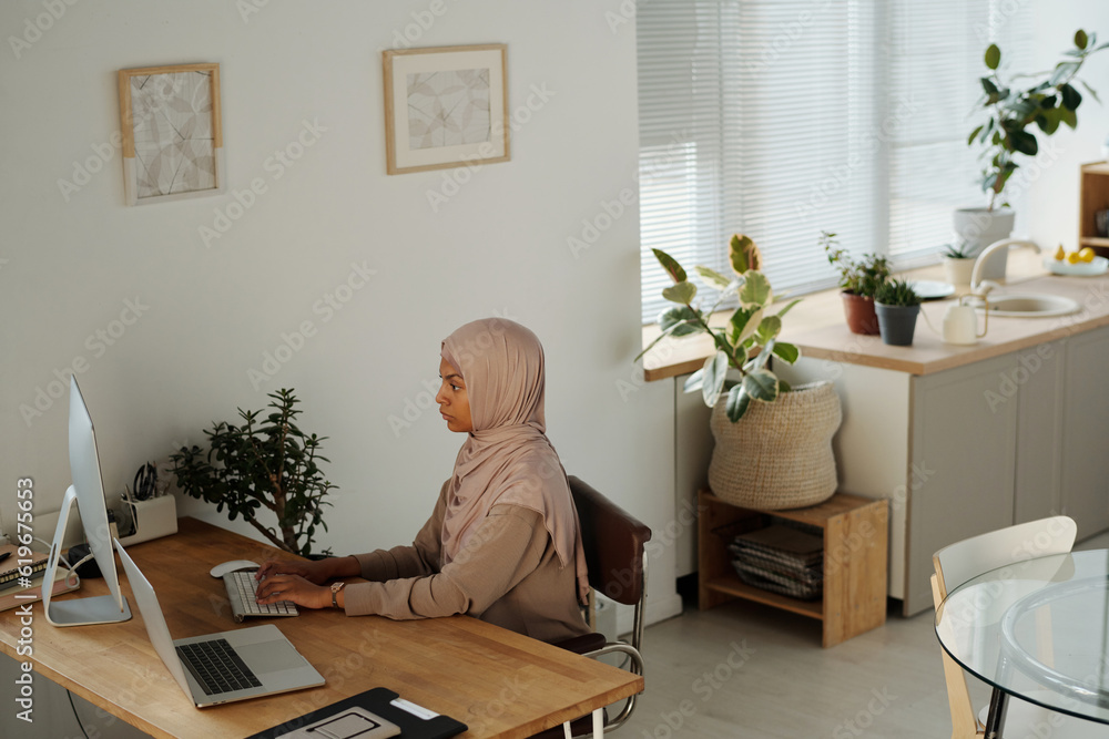Aufkleber Serious Muslim female IT manager in hijab sitting by desk in front of desktop computer monitor and decoding data or organizing work