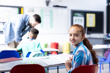 Obraz premium Smiling biracial schoolgirl sitting at desk in elementary school classroom, with copy space