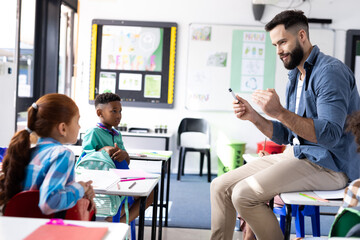 Diverse pupils and male teacher sitting on desk talking in elementary school classroom, copy space