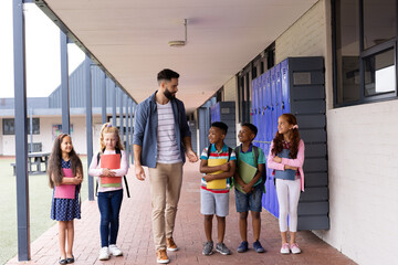 Obraz premium Diverse, happy male teacher and elementary schoolchildren talking in school corridor, copy space