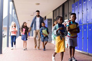 Fototapeta premium Diverse, happy male teacher and children walking together in elementary school corridor, copy space