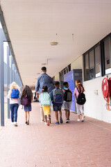 Vertical rear view of diverse male teacher and children in elementary school corridor, copy space