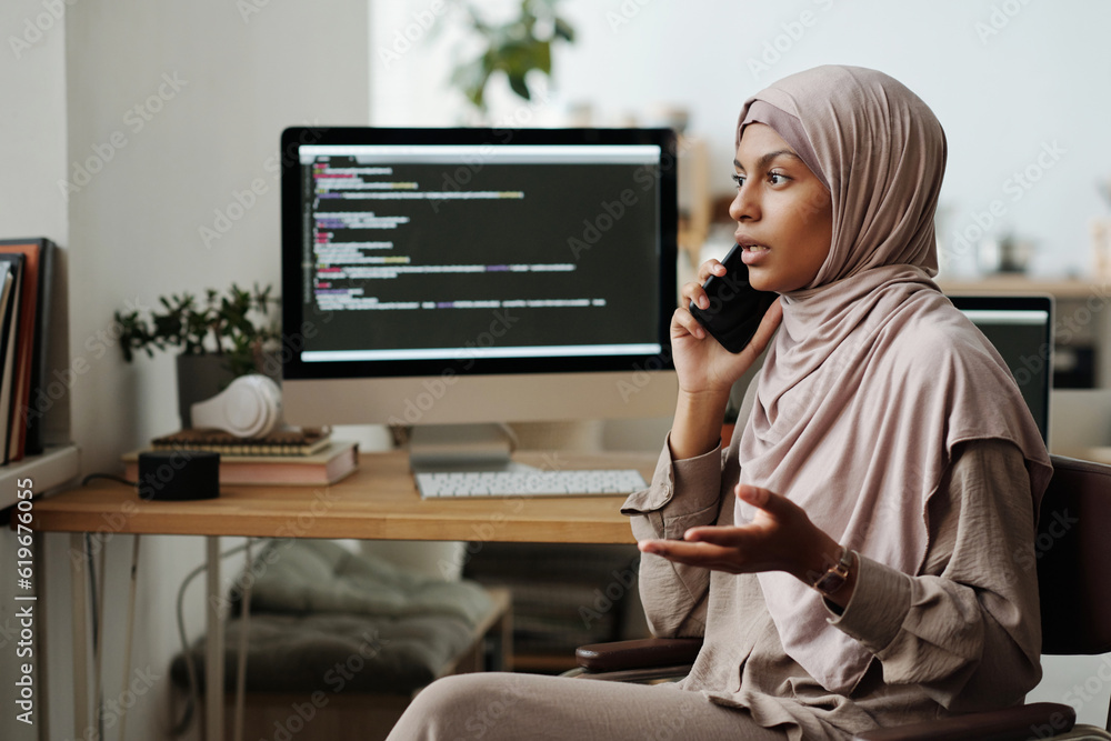 Aufkleber Young confident Muslim female programmer speaking on mobile phone while explaining something to colleague against computer screen