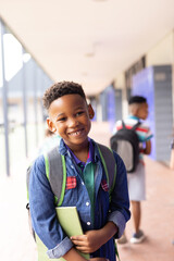 Vertical portrait of smiling african american boy in elementary school corridor, with copy space