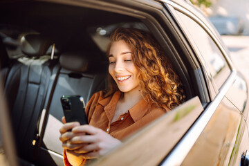 Smiling woman using smartphone while sitting in the back seat of a car. Young woman checks mail,...