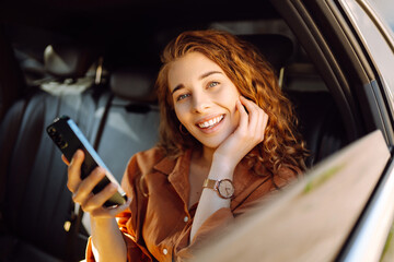 Smiling woman using smartphone while sitting in the back seat of a car. Young woman checks mail,...