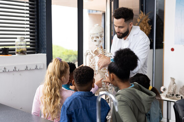 Diverse male teacher and elementary schoolchildren studying skeleton in biology class