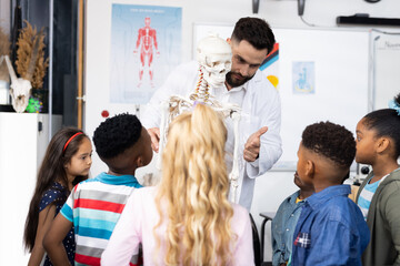 Diverse male teacher and elementary schoolchildren studying skeleton in biology class