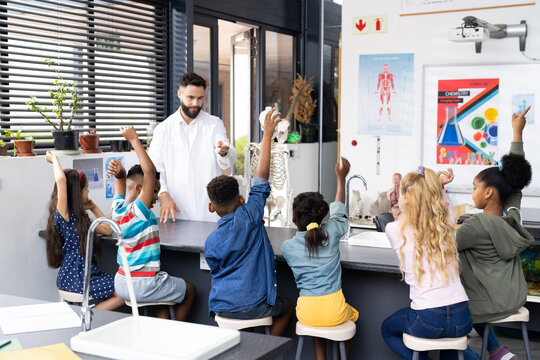 Diverse male teacher and elementary schoolchildren raising hands in biology class