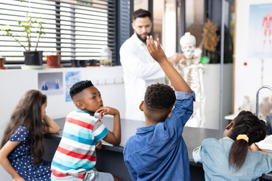 Diverse male teacher and elementary schoolchildren, one raising hand, in biology class