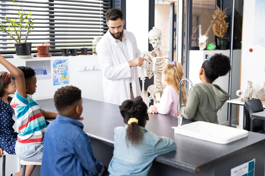 Diverse male teacher and elementary schoolchildren, one raising hand, in biology class